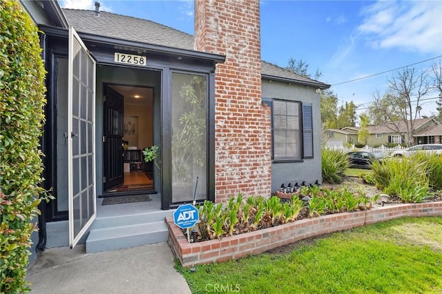 entrance to property featuring a shingled roof, stucco siding, a chimney, a yard, and brick siding