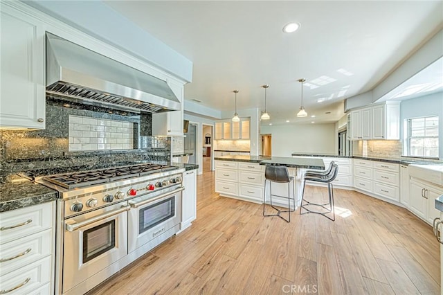 kitchen featuring white cabinets, hanging light fixtures, double oven range, wall chimney exhaust hood, and light hardwood / wood-style flooring