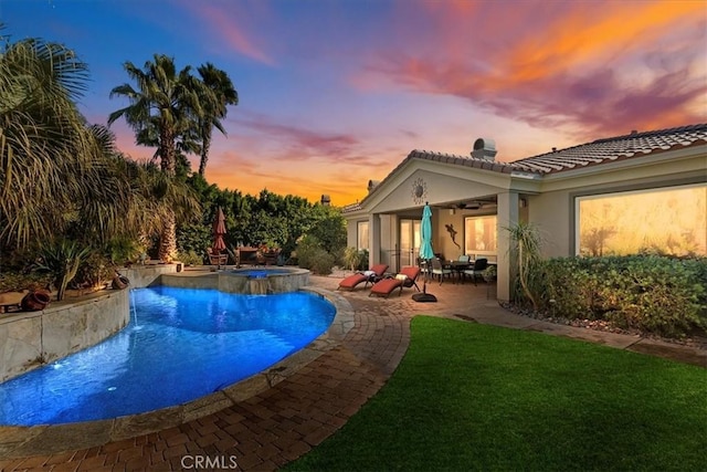 pool at dusk featuring ceiling fan, a patio, pool water feature, and an in ground hot tub