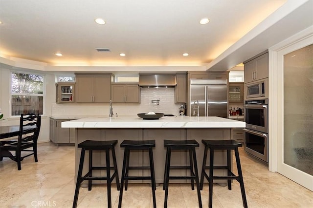 kitchen featuring a spacious island, wall chimney exhaust hood, tasteful backsplash, built in appliances, and a kitchen breakfast bar