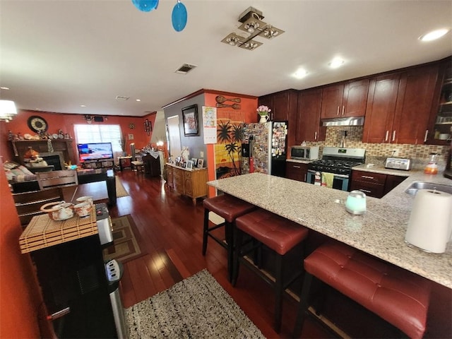 kitchen with appliances with stainless steel finishes, a breakfast bar area, backsplash, dark hardwood / wood-style flooring, and light stone counters