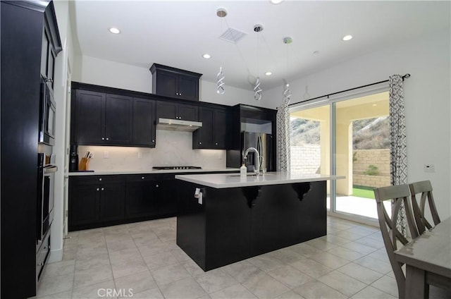 kitchen with under cabinet range hood, a sink, light countertops, dark cabinetry, and an island with sink