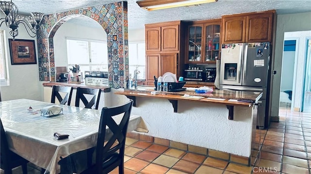 kitchen with a textured ceiling, stainless steel refrigerator with ice dispenser, glass insert cabinets, and brown cabinets