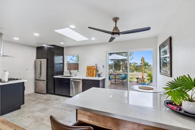 kitchen featuring wall chimney exhaust hood, sink, a skylight, appliances with stainless steel finishes, and ceiling fan