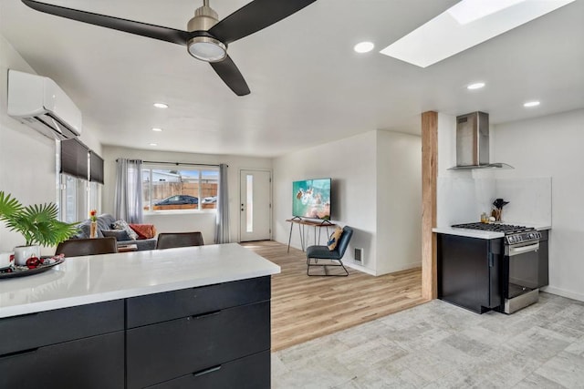 kitchen featuring wall chimney exhaust hood, a skylight, an AC wall unit, stainless steel range with gas stovetop, and ceiling fan