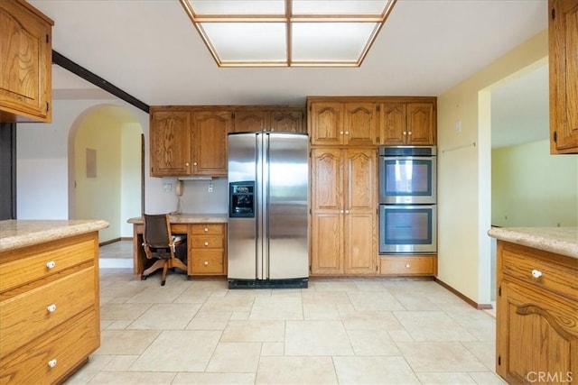 kitchen featuring arched walkways, brown cabinets, stainless steel appliances, light countertops, and built in desk