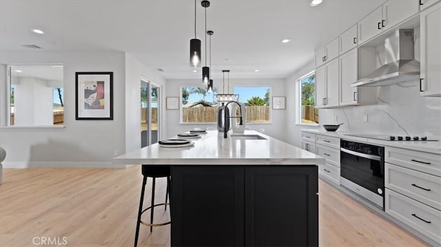 kitchen with white cabinetry, an island with sink, black electric cooktop, oven, and wall chimney exhaust hood