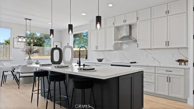 kitchen with a kitchen island with sink, light stone countertops, white cabinets, decorative light fixtures, and wall chimney exhaust hood