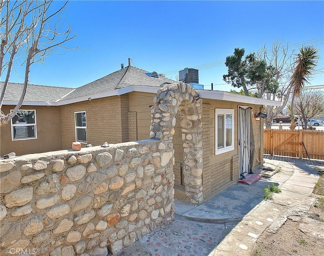 exterior space with central AC unit, brick siding, roof with shingles, and fence