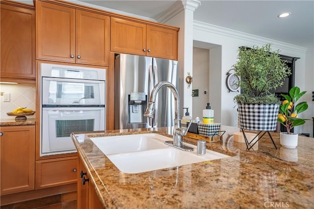 kitchen featuring sink, double oven, stainless steel refrigerator with ice dispenser, light stone counters, and ornamental molding