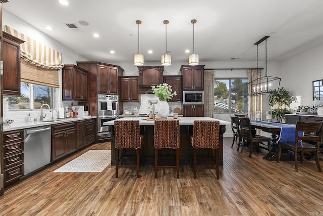 kitchen with dark wood-type flooring, decorative light fixtures, dark brown cabinets, appliances with stainless steel finishes, and a kitchen island with sink