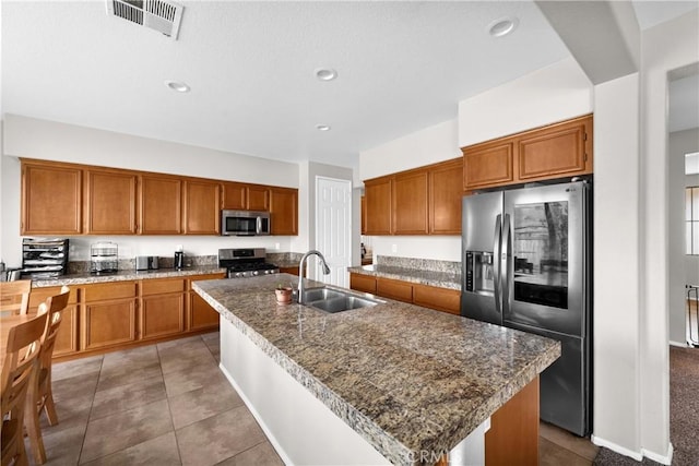 kitchen with an island with sink, brown cabinetry, stainless steel appliances, and a sink