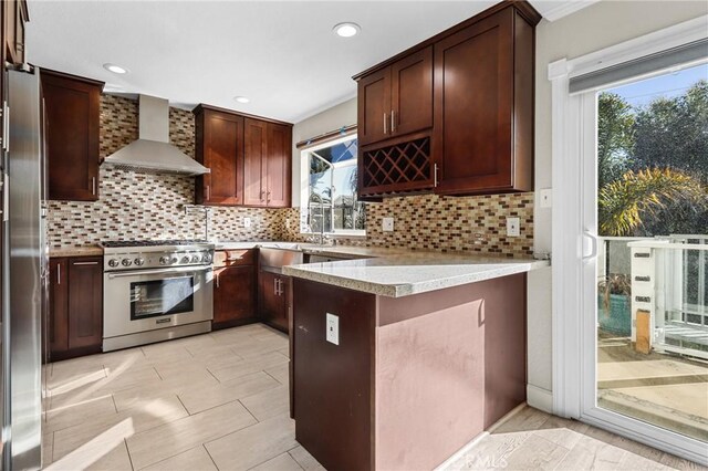 kitchen featuring wall chimney range hood, backsplash, kitchen peninsula, and appliances with stainless steel finishes