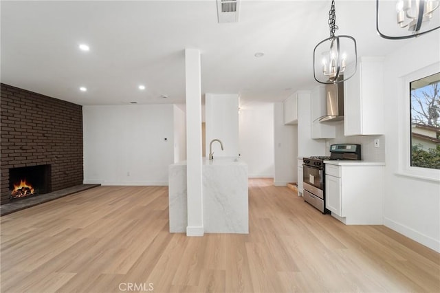 kitchen with white cabinetry, a brick fireplace, stainless steel range with gas cooktop, pendant lighting, and wall chimney range hood