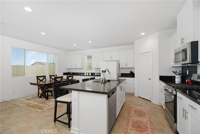 kitchen featuring an island with sink, a sink, stainless steel appliances, white cabinets, and dark countertops