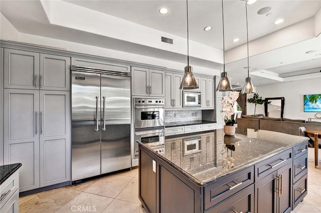 kitchen with pendant lighting, backsplash, built in appliances, a raised ceiling, and dark stone counters