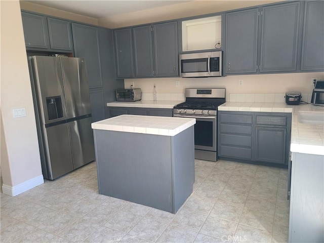 kitchen with stainless steel appliances, a kitchen island, baseboards, gray cabinets, and tile counters