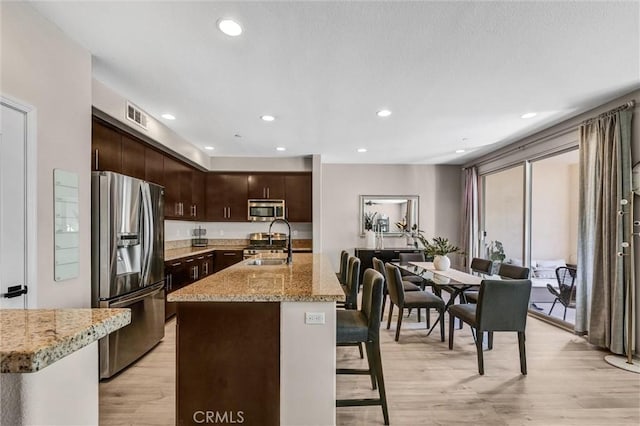 kitchen featuring light wood-style flooring, a sink, appliances with stainless steel finishes, light stone countertops, and an island with sink