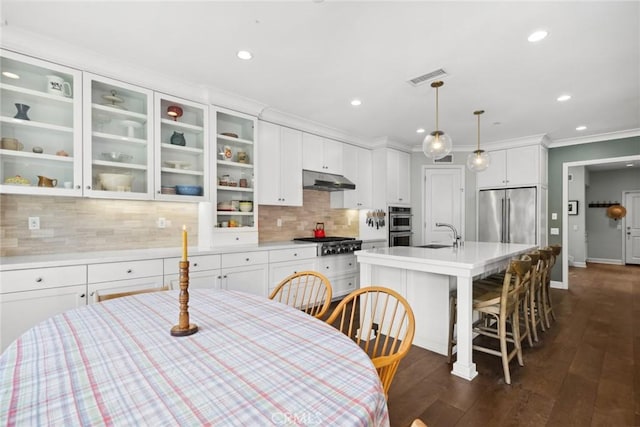 kitchen featuring dark wood-type flooring, appliances with stainless steel finishes, a kitchen island with sink, hanging light fixtures, and white cabinets