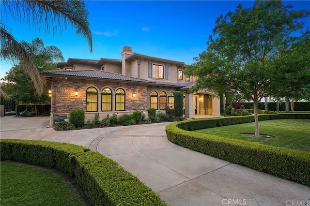 mediterranean / spanish home with a tile roof, a chimney, stone siding, driveway, and a front lawn