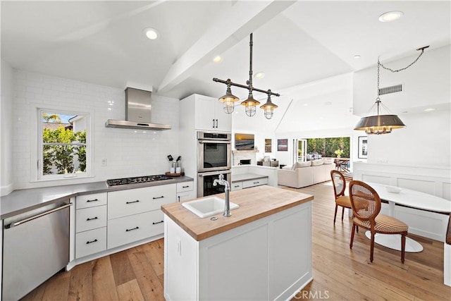 kitchen featuring a center island with sink, wall chimney range hood, pendant lighting, stainless steel appliances, and white cabinets