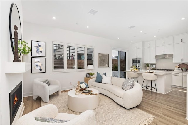 living room featuring light wood finished floors, a warm lit fireplace, visible vents, baseboards, and recessed lighting