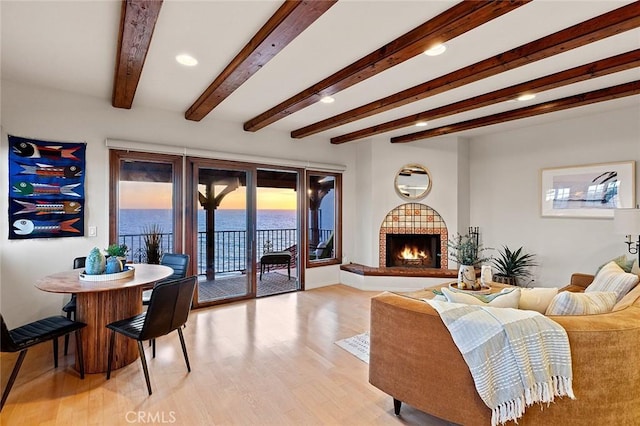 living room featuring a water view, a warm lit fireplace, light wood-style flooring, and beamed ceiling