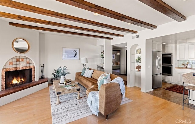 living area with a warm lit fireplace, beam ceiling, light wood-type flooring, and visible vents