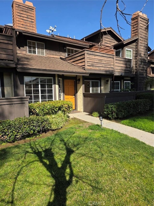 view of front facade with roof with shingles, a chimney, stucco siding, a balcony, and a front lawn