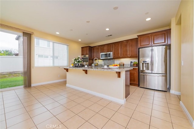 kitchen with a breakfast bar area, light tile patterned floors, light stone counters, stainless steel appliances, and a center island with sink