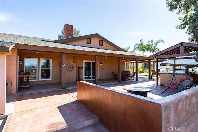 rear view of property featuring a chimney, roof with shingles, a gazebo, a patio area, and stucco siding