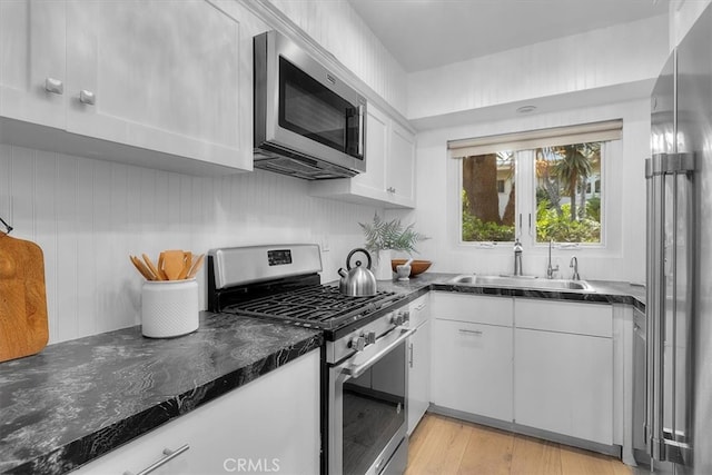 kitchen featuring stainless steel appliances, a sink, light wood-style floors, dark stone counters, and white cabinetry