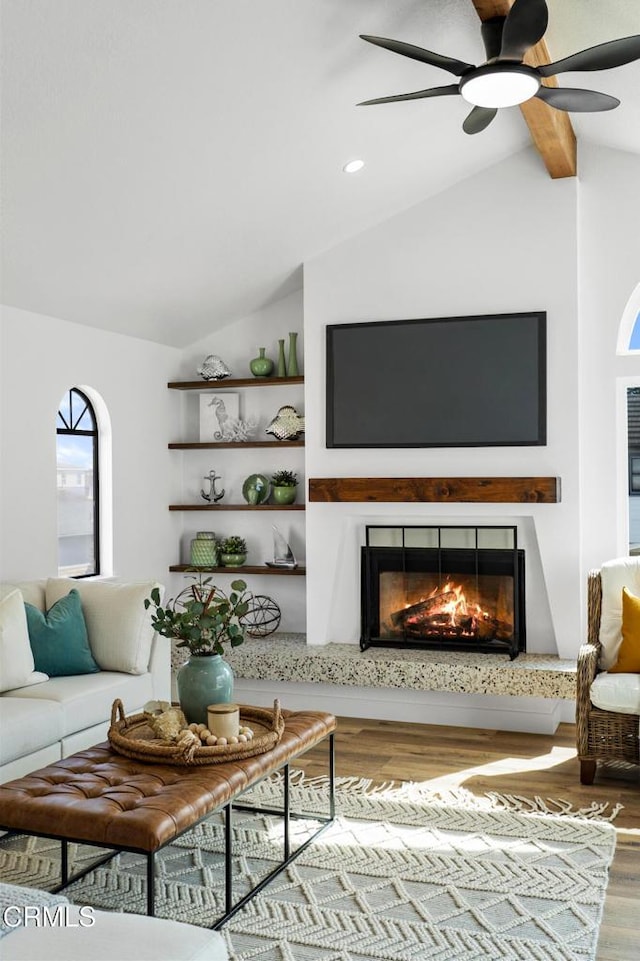 living area featuring vaulted ceiling with beams, ceiling fan, wood finished floors, and a glass covered fireplace