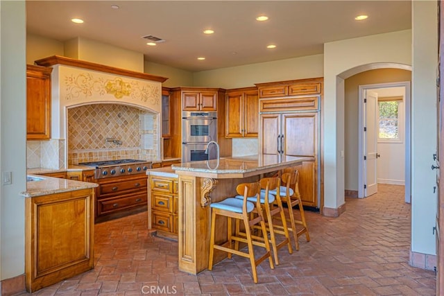 kitchen with appliances with stainless steel finishes, a kitchen island with sink, light stone counters, and a breakfast bar