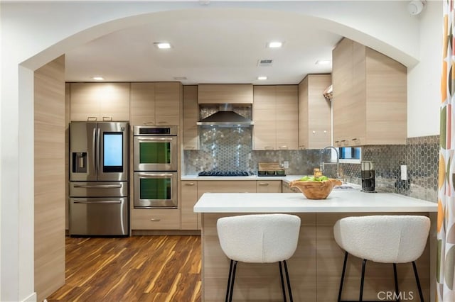 kitchen featuring dark hardwood / wood-style floors, light brown cabinetry, tasteful backsplash, stainless steel appliances, and wall chimney exhaust hood