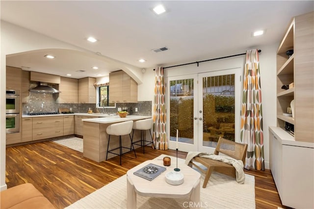 kitchen with wall chimney exhaust hood, light brown cabinets, kitchen peninsula, double oven, and black gas stovetop