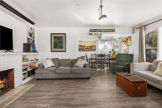 living room with a brick fireplace, dark wood-type flooring, and ceiling fan