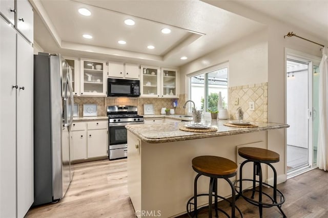 kitchen with a sink, light wood-style floors, stainless steel appliances, a peninsula, and a tray ceiling