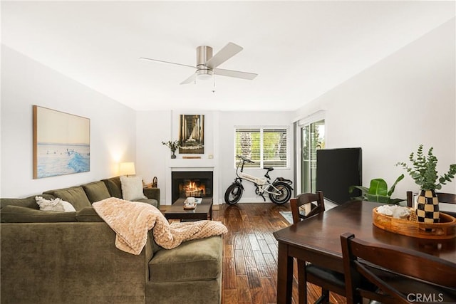 living room featuring a warm lit fireplace, ceiling fan, and dark wood-style flooring