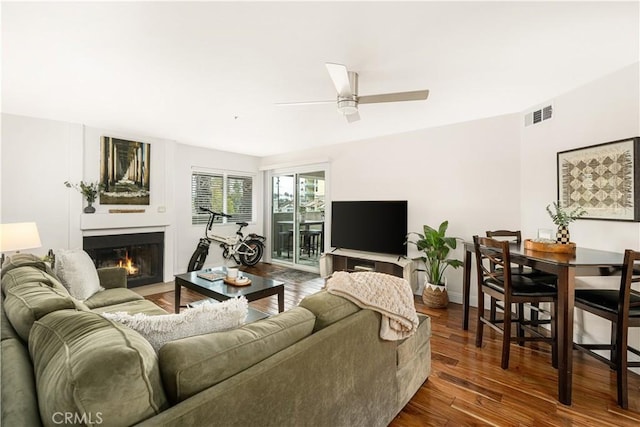 living area featuring visible vents, a ceiling fan, a glass covered fireplace, wood finished floors, and baseboards