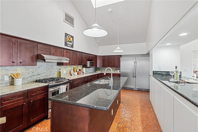 kitchen with sink, appliances with stainless steel finishes, dark stone countertops, an island with sink, and decorative light fixtures