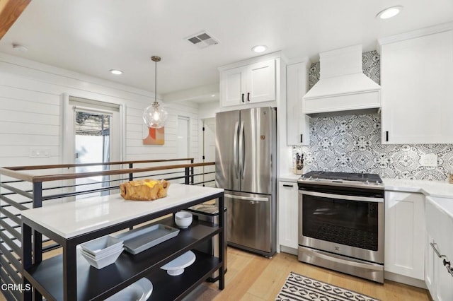 kitchen with light wood-type flooring, appliances with stainless steel finishes, custom range hood, pendant lighting, and white cabinets