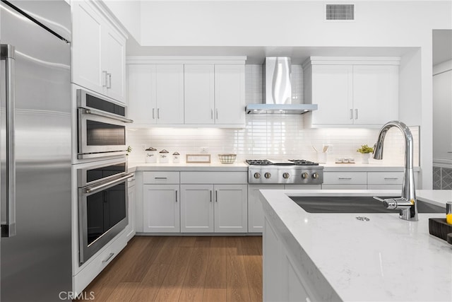 kitchen featuring tasteful backsplash, wall chimney range hood, stainless steel appliances, light stone countertops, and white cabinets