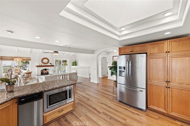 kitchen with stainless steel appliances, a fireplace, a raised ceiling, dark stone counters, and light wood-type flooring