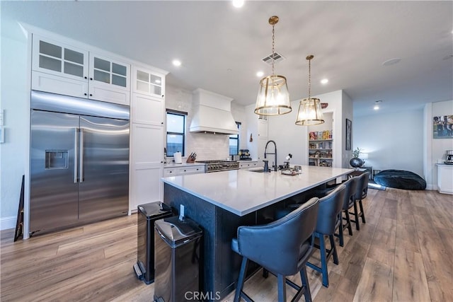 kitchen with pendant lighting, premium range hood, high quality appliances, white cabinets, and a center island with sink