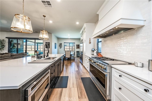 kitchen featuring premium range hood, decorative light fixtures, white cabinetry, an island with sink, and premium appliances