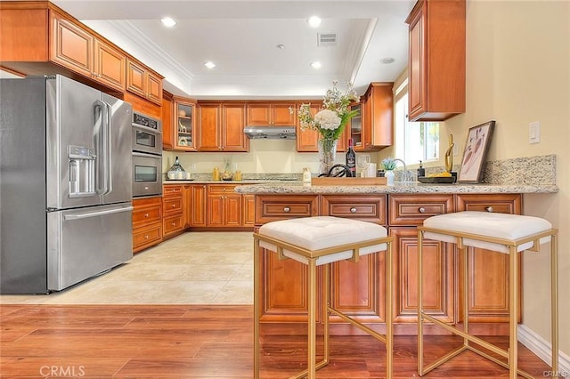 kitchen with appliances with stainless steel finishes, a kitchen breakfast bar, light hardwood / wood-style floors, crown molding, and light stone countertops