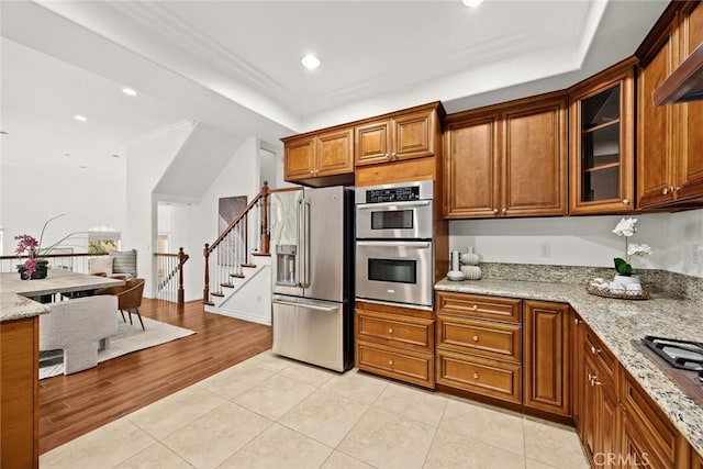 kitchen with light tile patterned flooring, glass insert cabinets, light stone countertops, brown cabinetry, and appliances with stainless steel finishes