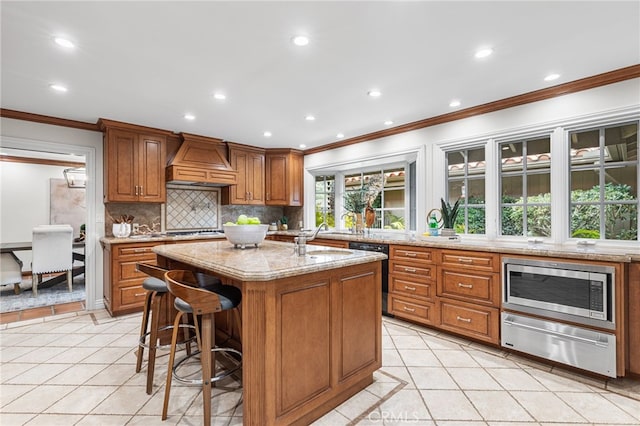 kitchen featuring tasteful backsplash, stainless steel microwave, custom range hood, light stone countertops, and a kitchen island with sink