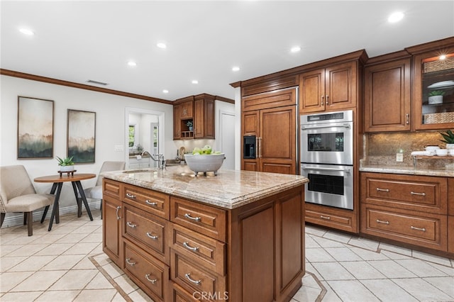 kitchen with crown molding, a kitchen island with sink, light tile patterned flooring, decorative backsplash, and stainless steel double oven